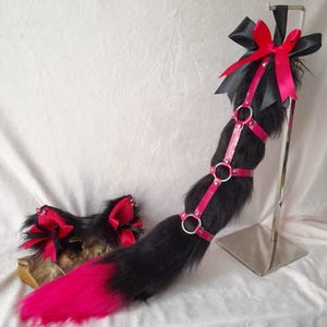 Może przedstawiać: Czarny i różowy ogon z imitacji futra z różową uprzężą i srebrnymi pierścieniami. Ogon jest eksponowany na srebrnym stojaku. Obecne są również pasujące czarno-różowe uszy z czerwonymi kokardkami.