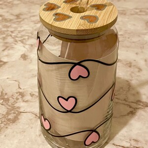 May include: A clear glass water bottle with a bamboo lid and a pink heart design. The lid has a straw hole and a straw is inserted through the hole. The bottle has a pink heart design with black outlines.