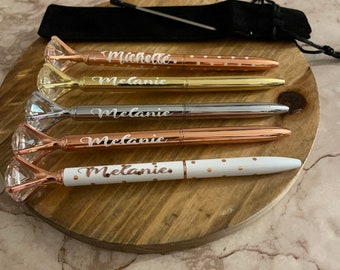 Puede incluir: Cinco bolígrafos personalizados con tapas en forma de diamante. Los bolígrafos son de oro, plata y oro rosa con lunares blancos. Los nombres "Michelle" y "Melanie" están grabados en los bolígrafos.