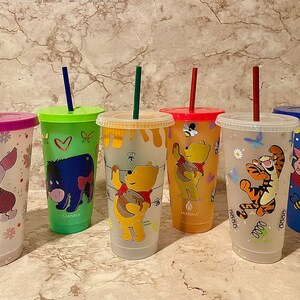 May include: Six clear plastic tumblers with colorful designs featuring characters from Winnie the Pooh. The tumblers have lids and straws. The designs include Tigger, Eeyore, Piglet, Winnie the Pooh, and a bee.