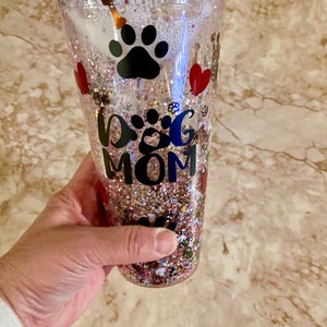 May include: Clear plastic tumbler with a pink, gold, and white glitter design. The tumbler has a black paw print, red hearts, and the text "Dog Mom".