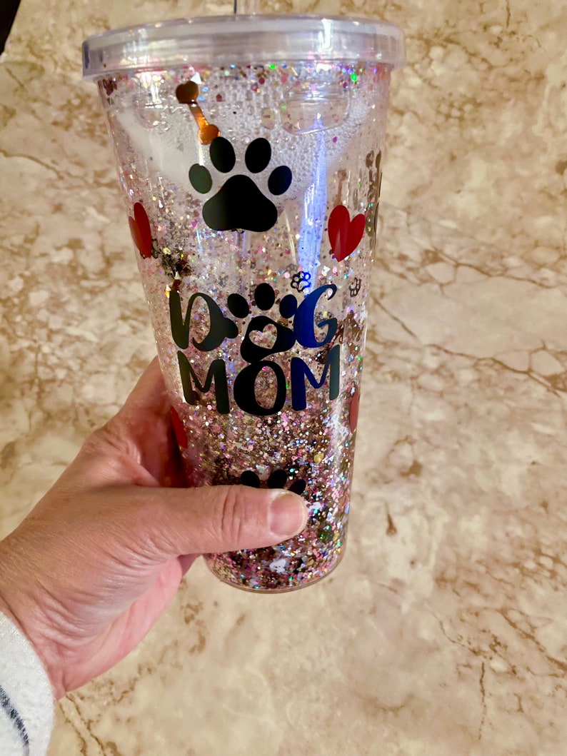 May include: Clear plastic tumbler with a pink, gold, and white glitter design. The tumbler has a black paw print, red hearts, and the text "Dog Mom".