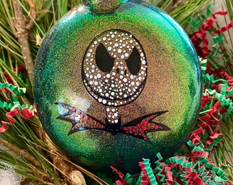 Puede incluir: Adorno navideño verde iridiscente con una cara de Jack Skellington en blanco y negro pintada en él. El adorno tiene detalles de purpurina roja.