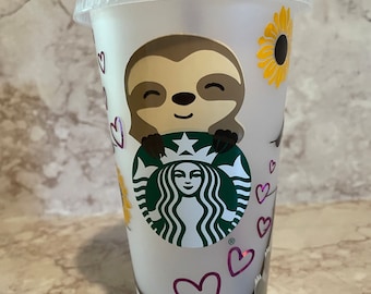 May include: A white reusable cup with a sloth holding a green Starbucks logo. The cup has pink hearts and yellow sunflowers around the sloth.