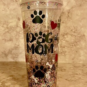 May include: Clear plastic tumbler with a pink, gold, and white glitter design. The tumbler has a black paw print, a red heart, and the text "Dog Mom" on it.