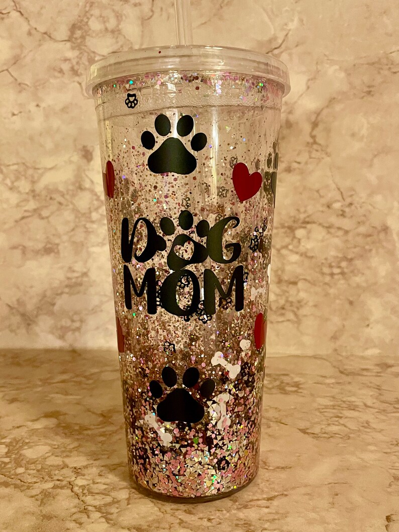 May include: Clear plastic tumbler with a pink, gold, and white glitter design. The tumbler has a black paw print, a red heart, and the text "Dog Mom" on it.