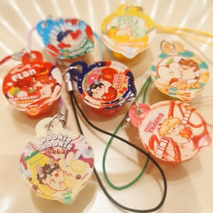 Puede incluir: Una colección de coloridos amuletos translúcidos con ilustraciones de dibujos animados y texto. Los amuletos presentan varios diseños, incluyendo una fresa, una manzana y un plátano, con cordones adjuntos. Los amuletos son de varias formas y tamaños.