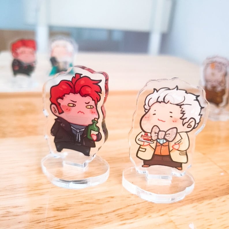 Op de afbeelding: Twee acryl karakterstandaards met cartoonfiguren. De figuur links is een roodharige man met een groene fles in zijn hand. De figuur rechts is een man met wit haar die een stuk taart vasthoudt. Beide figuren dragen kleding en hebben een cartoonachtige stijl.