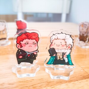 Op de afbeelding: Twee acryl karakterstandaards met cartoonfiguren. De figuur links is een man met rood haar, die een groen militair uniform draagt en een pistool vasthoudt. De figuur rechts is een man met wit haar, die een groen-blauw pak draagt met een witte kraag en een hoge hoed.