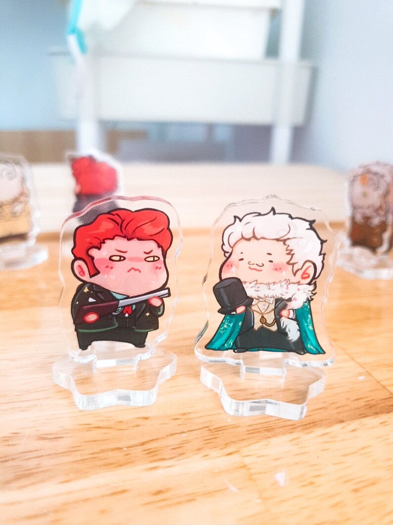 Op de afbeelding: Twee acryl karakterstandaards met cartoonfiguren. De figuur links is een man met rood haar, die een groen militair uniform draagt en een pistool vasthoudt. De figuur rechts is een man met wit haar, die een groen-blauw pak draagt met een witte kraag en een hoge hoed.