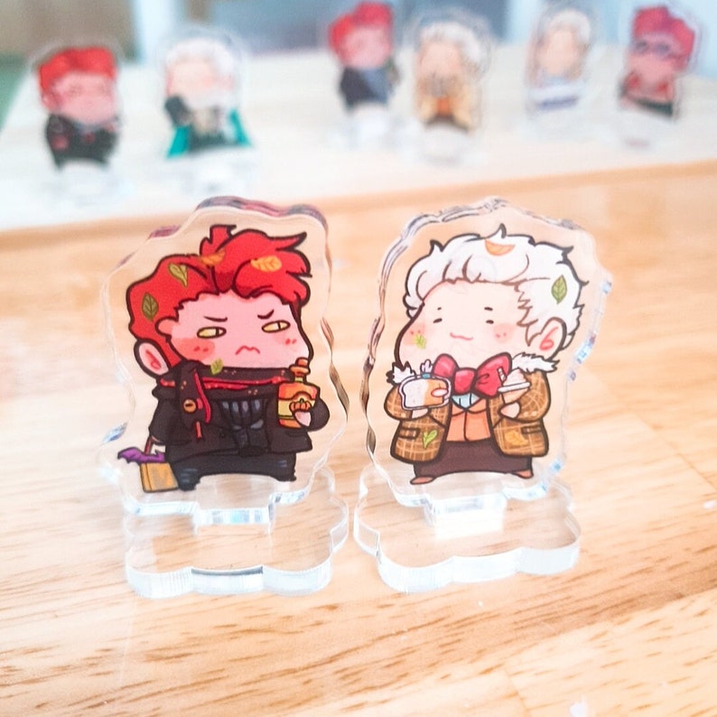 Op de afbeelding: Twee acryl karakterstandaards met cartoonfiguren. Het personage links is een roodharige man met een zwarte jas en een fles alcohol. Het personage rechts is een man met wit haar met een bruine jas en een kopje koffie. Beide personages hebben een cartoonachtige stijl met grote ogen en kleine monden.