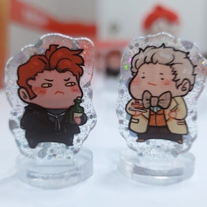 Op de afbeelding: Twee kleine, cartoonachtige acrylfiguren op heldere standaards. De figuur links heeft rood haar en houdt een fles vast. De figuur rechts heeft blond haar en houdt een stuk taart vast.