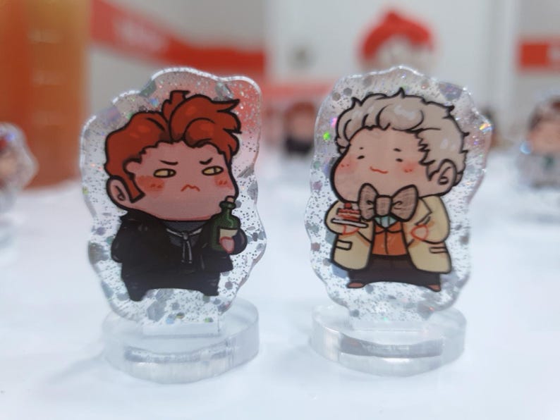 Op de afbeelding: Twee kleine, cartoonachtige acrylfiguren op heldere standaards. De figuur links heeft rood haar en houdt een fles vast. De figuur rechts heeft blond haar en houdt een stuk taart vast.