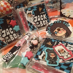 May include: A collection of merchandise featuring the text "JACK IN THE BOX" and the name "J-hope." Items include keychains with star-shaped charms, stickers, and a round pin. The designs incorporate a black and blue checkered pattern.