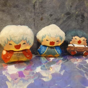 Könnte beinhalten: Drei handgefertigte Plüschpuppen mit lächelnden Gesichtern und farbenfrohen Outfits. Die Puppen haben weiße Haare mit blauen Akzenten, rote Münder und sind in roter, blauer und brauner Kleidung gekleidet. Sie werden auf einer Oberfläche mit schillernden Pailletten ausgestellt.