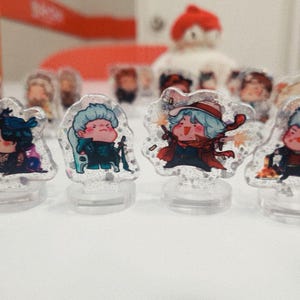 May include: Five colorful acrylic character figurines on clear stands. Each figurine is a different character with a unique design and pose. The figurines are on a white surface.