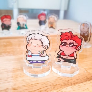 Op de afbeelding: Twee acryl figuurtjes op heldere standaards. Het figuurtje links is een wit haarig figuurtje met een rood-blauw badpak en een geel drankje. Het figuurtje rechts is een rood haarig figuurtje met een rood badpak en een zonnebril. Beide figuurtjes lachen.