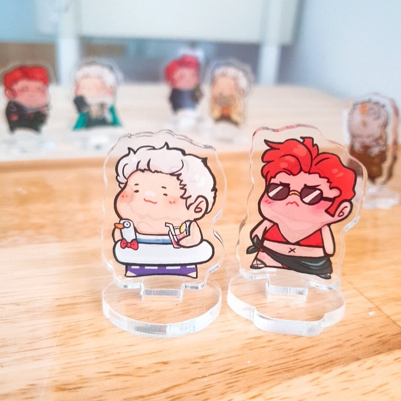 Op de afbeelding: Twee acryl figuurtjes op heldere standaards. Het figuurtje links is een wit haarig figuurtje met een rood-blauw badpak en een geel drankje. Het figuurtje rechts is een rood haarig figuurtje met een rood badpak en een zonnebril. Beide figuurtjes lachen.