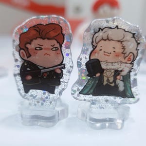 Op de afbeelding: Twee kleine, cartoonachtige figuurtjes op heldere acrylsokkels. Het figuurtje links is een roodharig personage met een serieuze uitdrukking, dat een pistool vasthoudt. Het figuurtje rechts is een blond personage met een vriendelijke uitdrukking, dat een groene jas draagt en een hoge hoed vasthoudt.