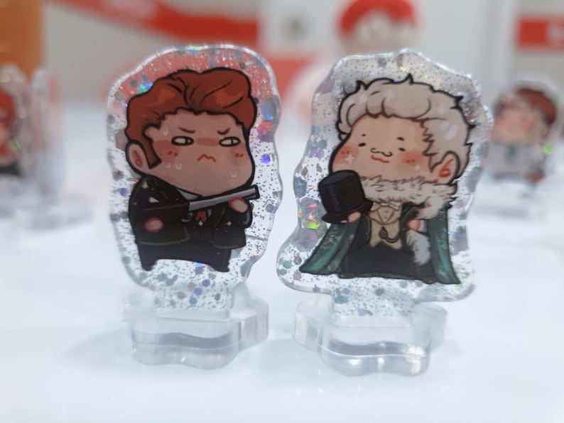 Op de afbeelding: Twee kleine, cartoonachtige figuurtjes op heldere acrylsokkels. Het figuurtje links is een roodharig personage met een serieuze uitdrukking, dat een pistool vasthoudt. Het figuurtje rechts is een blond personage met een vriendelijke uitdrukking, dat een groene jas draagt en een hoge hoed vasthoudt.