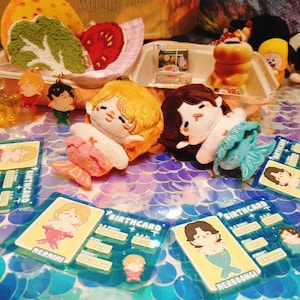 May include: A collection of mermaid-themed plush dolls and accessories. The dolls have yellow and brown hair, with mermaid tails. Birthcard-style cards with mermaid illustrations are also present, along with other small character figurines.