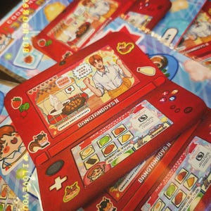 May include: A red handheld gaming console sticker featuring an anime-style character, the text BANGTANBOYS, and various food icons. The screen displays a cooking game interface with the text GOOD JOB, BABY and COOK WITH HOBA.