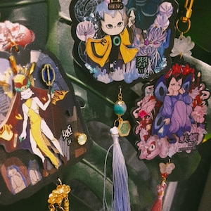 May include: A collection of colorful anime-style keychains with intricate designs. The keychains feature characters in various poses, with details like tassels, beads, and gold-colored hardware. The keychains are attached to a green leafy background.