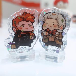 Op de afbeelding: Twee kleine, heldere harsfiguren met glitter ingebed in het hars. De figuren zijn van cartoonfiguren. Een personage is een roodharige persoon die een zwarte jas draagt en een kopje vasthoudt. Het andere personage is een blonde persoon die een bruine jas draagt en een kopje vasthoudt.