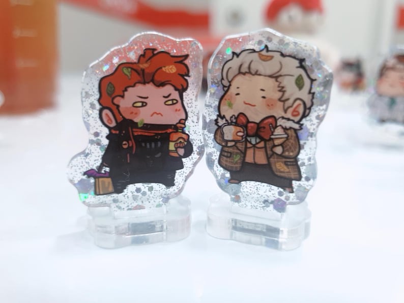 Op de afbeelding: Twee kleine, heldere harsfiguren met glitter ingebed in het hars. De figuren zijn van cartoonfiguren. Een personage is een roodharige persoon die een zwarte jas draagt en een kopje vasthoudt. Het andere personage is een blonde persoon die een bruine jas draagt en een kopje vasthoudt.