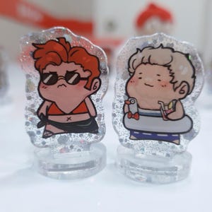 Op de afbeelding: Twee kleine, cartoonachtige figuurtjes op heldere standaards. Het figuurtje links is een persoon met rood haar die een zonnebril en een rood bikini-topje draagt. Het figuurtje rechts is een persoon met blond haar die een blauw-wit badpak draagt en een drankje en een kleine witte vogel vasthoudt.