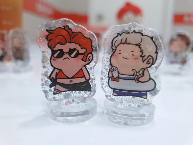 Op de afbeelding: Twee kleine, cartoonachtige figuurtjes op heldere standaards. Het figuurtje links is een persoon met rood haar die een zonnebril en een rood bikini-topje draagt. Het figuurtje rechts is een persoon met blond haar die een blauw-wit badpak draagt en een drankje en een kleine witte vogel vasthoudt.