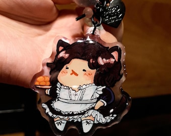 May include: A clear acrylic keychain featuring a cartoon character dressed in a maid outfit with cat ears, holding a tray of pastries. The keychain has a black chain, a gray pom-pom, and a heart-shaped charm.