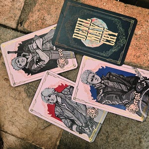 Könnte beinhalten: Ein Set aus vier rechteckigen Spielkarten mit Devil May Cry-Branding. Eine Karte ist schwarz mit goldenem Text, während die anderen drei illustrierte Charaktere namens Nero, Dante und Vergil vor rosa und blauen Hintergründen zeigen.