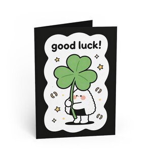 Może przedstawiać: Czarna kartka z pozdrowieniami z napisem "good luck!" w kolorze białym. Karta przedstawia postać z kreskówki trzymającą zieloną czterolistną koniczynę. Postać jest biała z czarnym strojem i różowym rumieńcem.