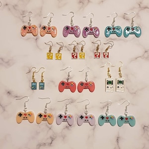May include: Assortment of colourful video game themed earrings. The earrings feature various designs including game controllers, question blocks, and game consoles.