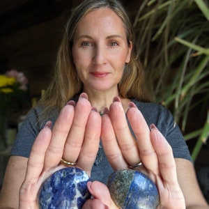 May include: Two smooth, oval-shaped, polished lapis lazuli stones, one with a darker blue hue and the other with a lighter blue hue, are held in the palms of a person's hands.