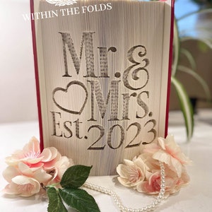 Op de afbeelding: Een wit boek met de woorden "Mr. & Mrs. Est. 2023" in de kaft gegraveerd. Het boek is dicht en staat rechtop op een wit oppervlak. Er liggen roze bloemen en een parelketting voor het boek.