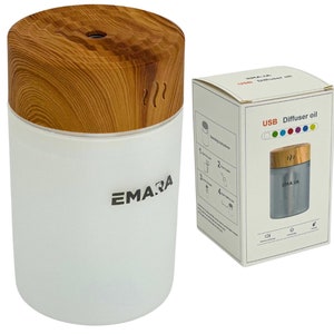 Op de afbeelding: Een witte en houten USB-etherische olieverdamper met een houten bovenkant en een witte basis. De diffuser heeft een zilveren label met het woord "EMARA" erop. De diffuser staat op een wit oppervlak. De doos voor de diffuser is wit met een zilveren label waarop "USB Diffuser oil" staat en een afbeelding van de diffuser.