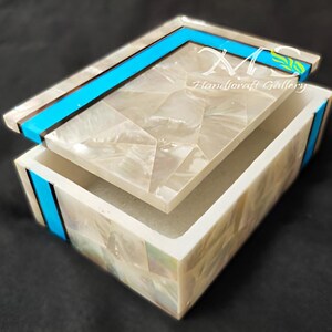 May include: A white mother of pearl box with a turquoise and black trim. The box is open and the lid is resting on top of the box. The box is made of inlaid mother of pearl and has a polished finish.
