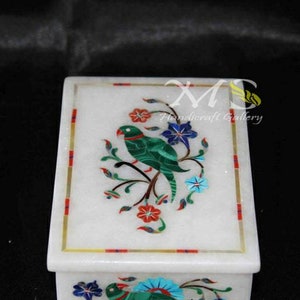 Puede incluir: Caja de mármol blanco con un pájaro verde y un diseño floral incrustado con piedras de colores. La caja tiene un borde dorado.