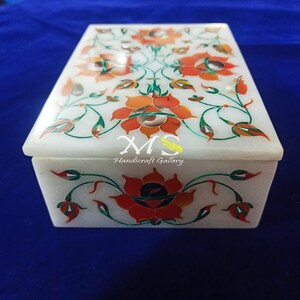 Puede incluir: Caja de mármol blanco con un diseño floral incrustado con piedras rojas, naranjas, verdes y doradas. La caja está decorada con un patrón floral y el texto "MS Handicraft Gallery".