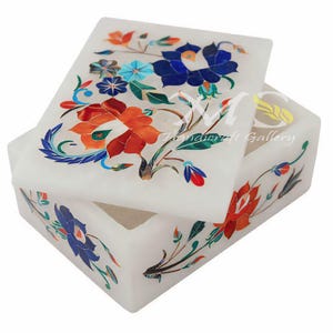 Puede incluir: Caja de mármol blanco con un diseño floral incrustado con piedras de colores. La tapa está abierta y la caja está decorada con flores azules, rojas, verdes y naranjas.