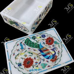 Puede incluir: Una caja y tapa decorativas de mármol blanco con intrincados diseños florales y de aves incrustados en tonos rojos, azules, verdes y amarillos. La caja rectangular y la tapa muestran una artesanía detallada.