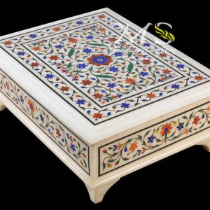 May include: A white marble box with intricate floral designs inlaid with colorful stones. The box has a square shape and a raised lid.