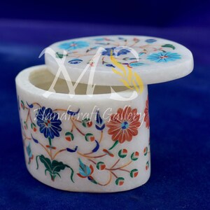 May include: White oval-shaped marble trinket box with a floral design inlaid with colorful stones. The lid is open and the box is empty.