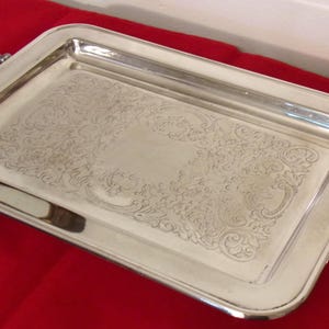 May include: A rectangular silver serving tray with ornate handles and a decorative floral pattern. The tray is set on a red surface, showcasing its polished finish and elegant design, suitable for serving food or displaying items.