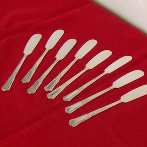 May include: An assortment of eight silver butter spreaders arranged on a vibrant red surface. Each spreader features a wide, rounded blade and a decorative handle. The blades are approximately 5 inches long.