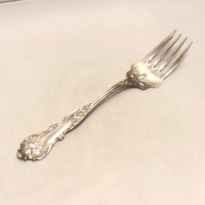 May include: A silver-toned metal fork featuring an ornate, embossed floral and scrollwork pattern along the handle and the base of the tines. The four-tined utensil has a polished, reflective surface with intricate detailing throughout the stem.