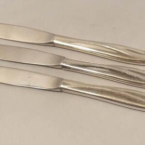 Puede incluir: Tres cuchillos plateados con hojas curvas y mangos ornamentados. Los cuchillos est&aacute;n dispuestos en diagonal, mostrando sus superficies pulidas y detalles decorativos. Un juego de cubiertos cl&aacute;sico.