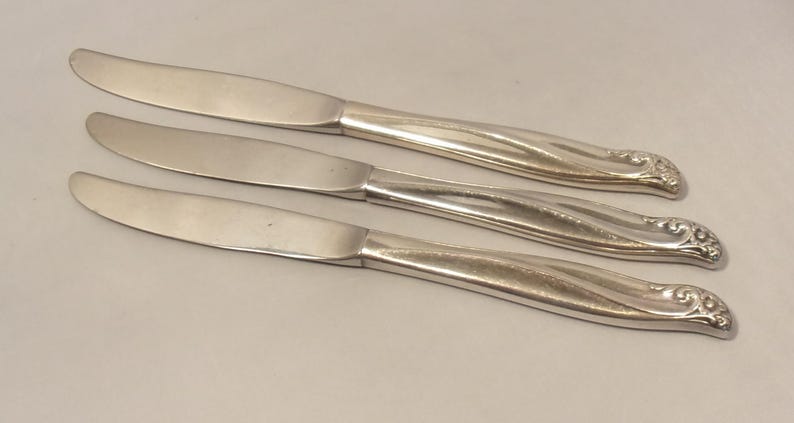 Puede incluir: Tres cuchillos plateados con hojas curvas y mangos ornamentados. Los cuchillos est&aacute;n dispuestos en diagonal, mostrando sus superficies pulidas y detalles decorativos. Un juego de cubiertos cl&aacute;sico.